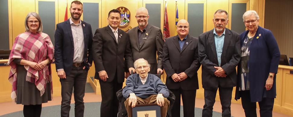 An older man (Trelle Morrow) surrounded by city councilors in Council Chambers.